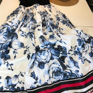 May include: A black sleeveless top and a white maxi dress with a blue floral pattern. The dress has a wide skirt with a border of black, red, and white stripes. A straw sun hat is also visible.