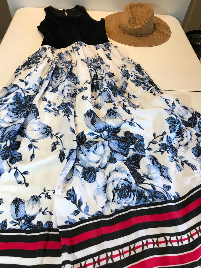 May include: A black sleeveless top and a white maxi dress with a blue floral pattern. The dress has a wide skirt with a border of black, red, and white stripes. A straw sun hat is also visible.
