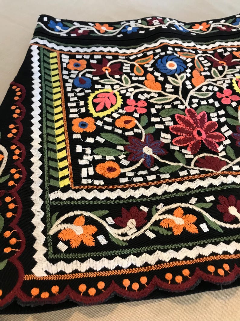 Puede incluir: Una falda negra con bordado floral de colores. El bordado presenta una variedad de flores, hojas y patrones geom&eacute;tricos en tonos de rojo, naranja, amarillo, verde, azul y blanco. La falda tiene un dobladillo festoneado.