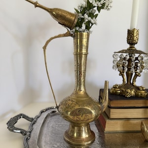 May include: A tall, ornate brass ewer with a long, slender neck, a bulbous base, and an attached lid, featuring intricate etched floral and geometric patterns. The metal vessel has a curved handle and a spout, with white floral stems inside.