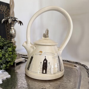 May include: A cream-colored ceramic teapot with a high, arched handle and a lid topped with a small building sculpture. The body features hand-painted illustrations of city skyscrapers, a church, and a yellow sun against light blue clouds.
