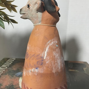 May include: A terracotta-coloured clay figurine featuring a dual-faced design with a ram-like head on one side and a human profile on the other. The conical base is decorated with painted white and dark red patterns, and a thin string encircles the neck.