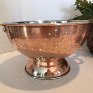 May include: A copper colander with a stainless steel base and handles. The colander has a hammered finish and a pattern of small holes.