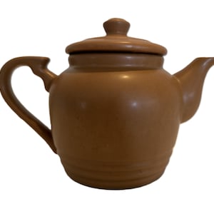 May include: A brown ceramic teapot with a lid, handle, and spout. The teapot has a rounded body and a small knob on the lid. The handle is curved, and the spout is angled. The teapot is a solid brown color.