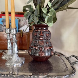 Może przedstawiać: Ciemnobrązowy ceramiczny wazon z białymi, wygrawerowanymi geometrycznymi wzorami i stylizowanym motywem twarzy, w towarzystwie dwóch srebrzystych metalowych świeczników z żółtymi świecami stożkowymi, na prostokątnej srebrzystej metalowej tacy.
