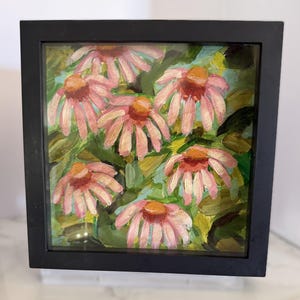May include: A square painting of pink coneflower blossoms with orange centers and green foliage, encased in a simple black frame. The artwork features visible brushstrokes and a layered application of paint, capturing the flowers in a garden setting.