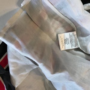 May include: Close-up of a white garment with a black and red striped accent. A small tag with text is visible. The garment appears to be made of a lightweight, semi-sheer fabric.