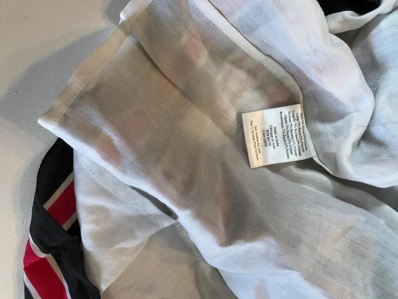 May include: Close-up of a white garment with a black and red striped accent. A small tag with text is visible. The garment appears to be made of a lightweight, semi-sheer fabric.