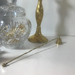 May include: A clear, cut-glass jar with a lid, a brass candlestick with a twisted design, and a brass bell with a long, twisted handle. The objects are arranged on a white surface.