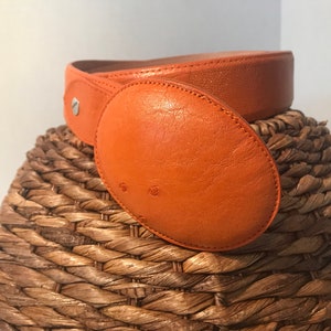 Op de afbeelding: Een feloranje leren riem met een grote ovale gesp. De riem ligt op een geweven mand.