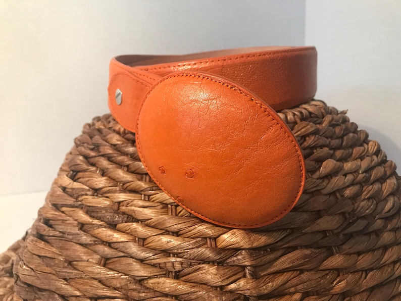 Op de afbeelding: Een feloranje leren riem met een grote ovale gesp. De riem ligt op een geweven mand.