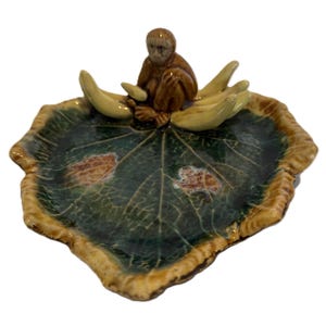 May include: A ceramic leaf-shaped dish with a monkey figurine and banana-shaped accents. The dish features a dark green and brown color scheme with a textured leaf pattern. The monkey is brown, and the bananas are a light yellow.