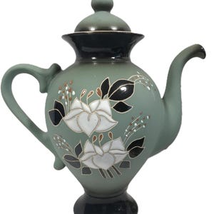 May include: A sage green ceramic teapot with a black base and accents. The teapot features a floral design with white flowers and black leaves. The lid has a finial. The handle and spout are curved.