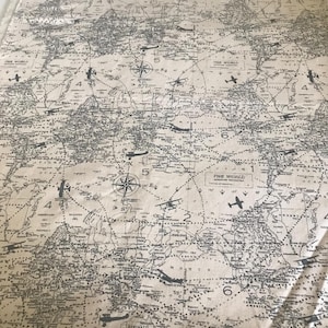 Op de afbeelding: Een beige stof met een zwart-witte print van een wereldkaart met vliegtuigen en kompasrozen. De tekst "THE WORLD" staat meerdere keren op de kaart.