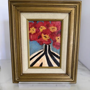 May include: An oil painting in a gold frame with a beige fabric mat. The artwork depicts a vase with black and white vertical stripes containing a bouquet of red flowers with yellow centers against a light blue background.