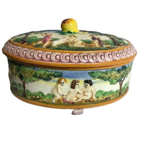 May include: A ceramic trinket box with a lid, featuring a painted pastoral scene with figures and trees. The lid has a yellow knob. The box is decorated with a repeating pattern around the rim and a scene of figures.