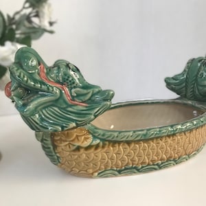May include: A ceramic dragon boat dish featuring a glossy green and tan finish. The vessel is shaped like a boat with detailed dragon heads at both ends, showcasing a textured scale pattern along the sides and a smooth, light-colored interior basin.