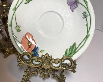 May include: A white porcelain saucer with a floral design featuring orange, purple, and green flowers. The saucer is on a gold metal stand.