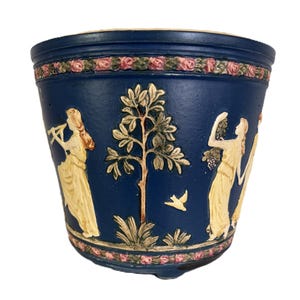 May include: A dark blue ceramic planter with a decorative relief design. The planter features classical figures, a tree, and a bird. A band of pink roses and green leaves encircles the top and bottom of the planter.