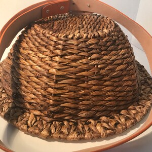 Op de afbeelding: Een geweven strooien mand met een bruine leren riem. De mand is rond en heeft een brede opening. De riem is met metalen klinknagels aan de mand bevestigd.