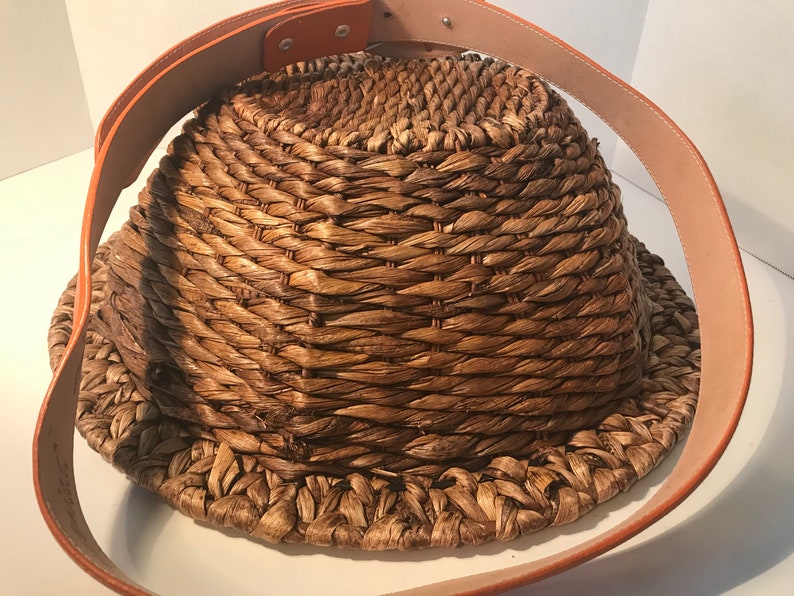 Op de afbeelding: Een geweven strooien mand met een bruine leren riem. De mand is rond en heeft een brede opening. De riem is met metalen klinknagels aan de mand bevestigd.