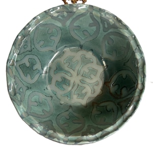 Może przedstawiać: Turkusowa ceramiczna miska z dekoracyjnym wzorem i mosiężnym uchwytem. Miska ma falisty brzeg i centralny kwiatowy wzór. Wzór jest w jaśniejszym odcieniu turkusu.