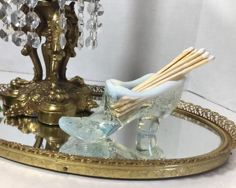 May include: A glass slipper match holder with a white opalescent finish. The holder is filled with wooden matches and sits on a gold mirrored tray with a decorative border.