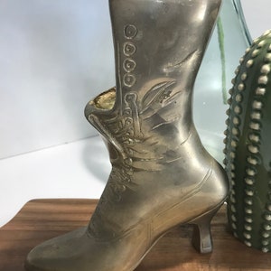 May include: A silver metal boot-shaped vase with a high heel. The boot has a decorative design and is sitting on a wooden surface.