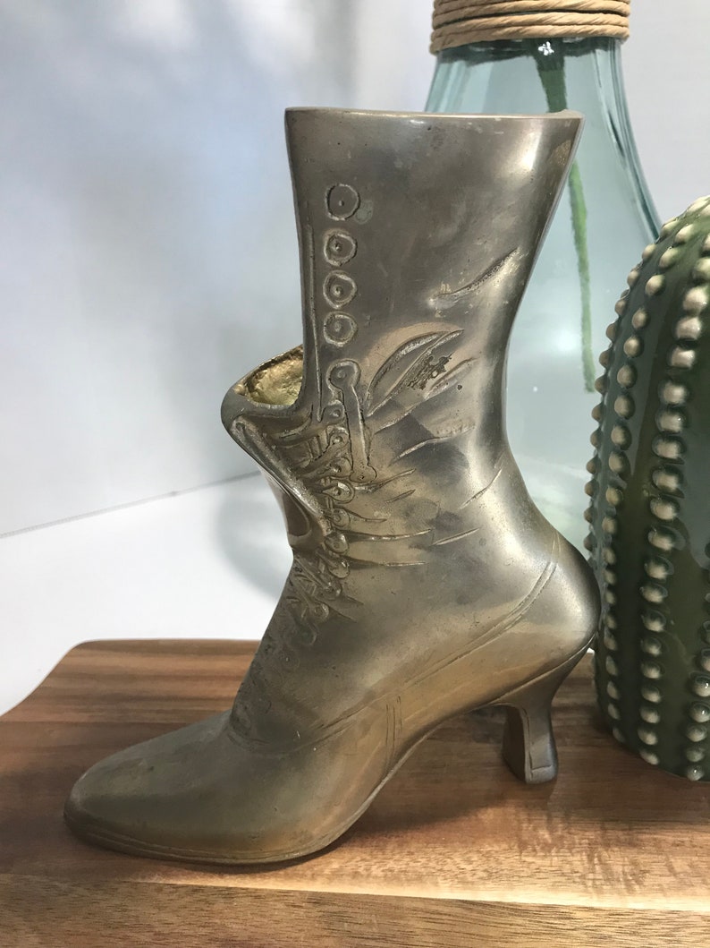 May include: A silver metal boot-shaped vase with a high heel. The boot has a decorative design and is sitting on a wooden surface.