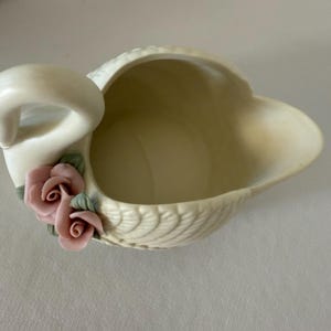 May include: Cream-colored swan-shaped ceramic dish with a detailed feather design. The swan features a curved neck and is adorned with two pink rose accents with green leaves. A decorative piece for the home.