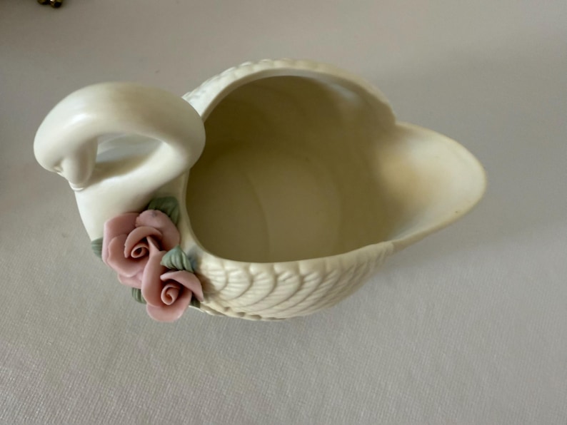 May include: Cream-colored swan-shaped ceramic dish with a detailed feather design. The swan features a curved neck and is adorned with two pink rose accents with green leaves. A decorative piece for the home.