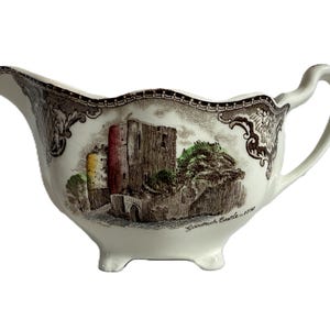 May include: Cream-colored gravy boat with a brown transferware design featuring a castle scene. The boat has a spout, handle, and three small feet. The rim is decorated with a patterned border. The text "Goodrich Castle - 1790" is visible.