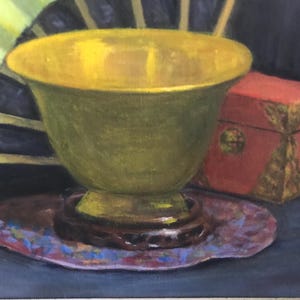 May include: A green ceramic bowl with a gold rim sits on a purple and blue patterned coaster. A red box with gold accents is behind the bowl.