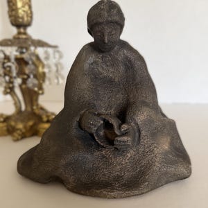 May include: A bronze-coloured sculpture of a seated figure, possibly a monk or a person in meditation. The figure is draped in a robe and holds a small object. The sculpture has a textured surface and is placed on a white surface.