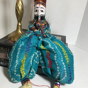 May include: A traditional Indian puppet with a painted face, wearing a colorful turban and a blue and white patterned outfit. The puppet is sitting on a book and has a red string attached to its hand.