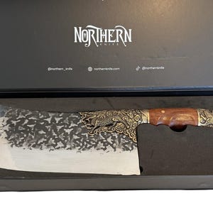May include: A cleaver knife with a wooden handle and ornate metal detailing, presented in a black box. The blade has a textured pattern. The box has the text "NORTHERN KNIVES".