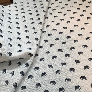 Può includere: Tessuto bianco trapuntato con un motivo ripetitivo di elefanti neri. Il tessuto è piegato a metà, mostrando il motivo su entrambi i lati.