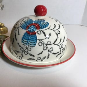 May include: A white ceramic butter dish with a domed lid, featuring a floral design in blue, red, and black. The lid is topped with a red knob. The dish sits on a matching plate with a red rim.