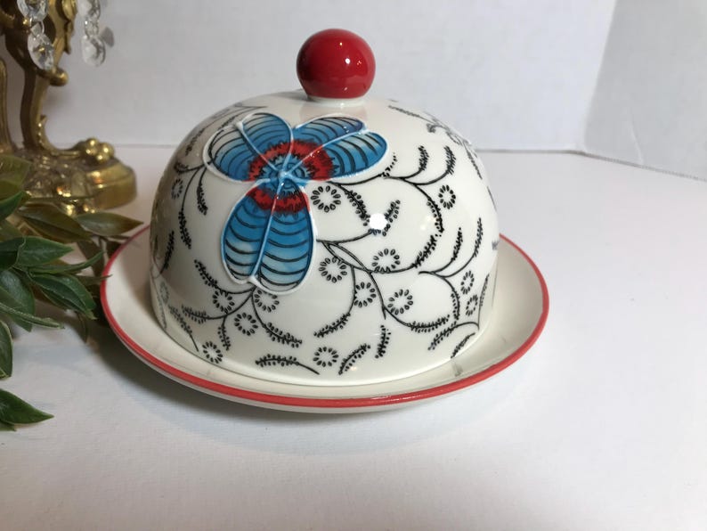 May include: A white ceramic butter dish with a domed lid, featuring a floral design in blue, red, and black. The lid is topped with a red knob. The dish sits on a matching plate with a red rim.
