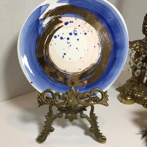 May include: A white ceramic plate with a blue and gold abstract design. The plate is on a gold metal stand.