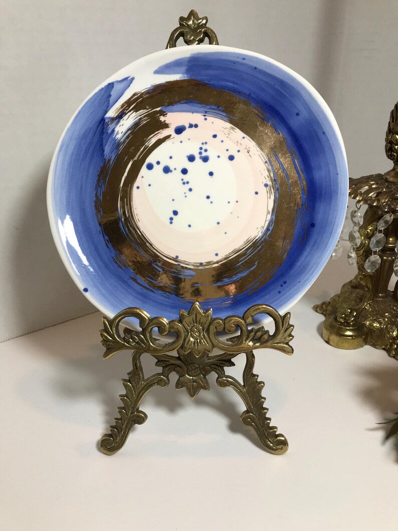 May include: A white ceramic plate with a blue and gold abstract design. The plate is on a gold metal stand.
