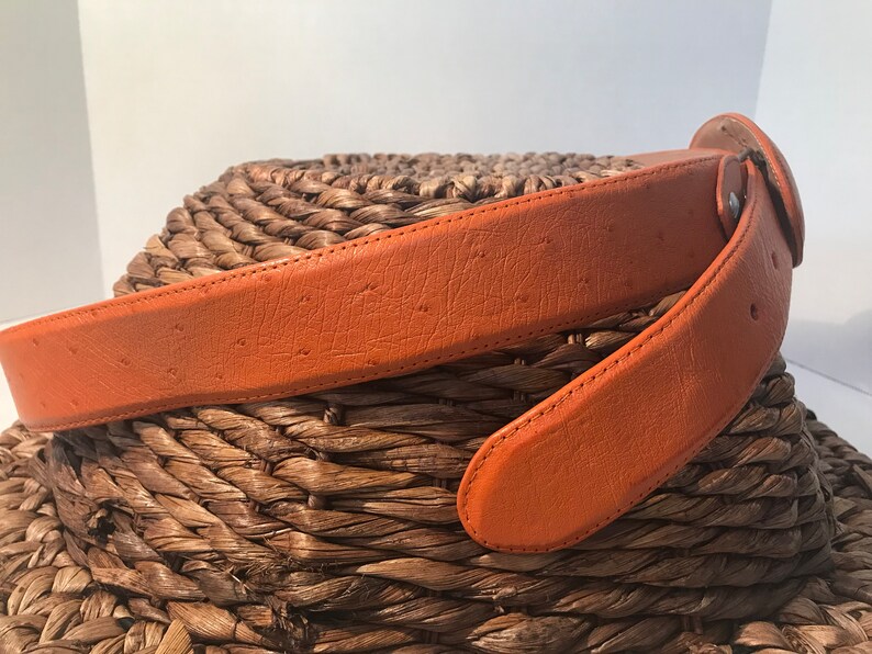 Op de afbeelding: Een bruine leren riem met een gestructureerd oppervlak. De riem is dubbelgevouwen en geplaatst op een geweven oppervlak.