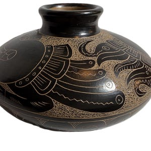 May include: A black ceramic vase with a rounded body and a short, cylindrical neck. The vase features intricate etched designs in a lighter colour, depicting stylized bird and geometric patterns. The overall aesthetic is reminiscent of ancient pottery.