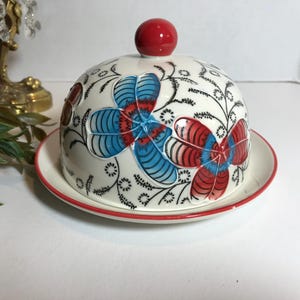 May include: A white ceramic butter dish with a domed lid and a matching plate. The lid is adorned with blue and red floral designs and a red knob. The plate and lid have a red trim. Decorative item.