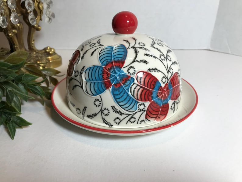 May include: A white ceramic butter dish with a domed lid and a matching plate. The lid is adorned with blue and red floral designs and a red knob. The plate and lid have a red trim. Decorative item.