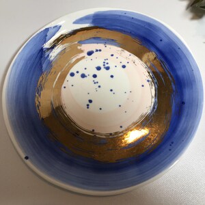 May include: A white ceramic plate with a blue and gold design. The plate has a circular design with a gold center and blue outer ring. The center of the plate has a pink and white design with blue dots.
