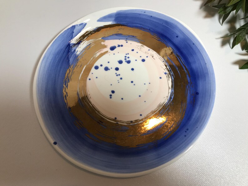 May include: A white ceramic plate with a blue and gold design. The plate has a circular design with a gold center and blue outer ring. The center of the plate has a pink and white design with blue dots.