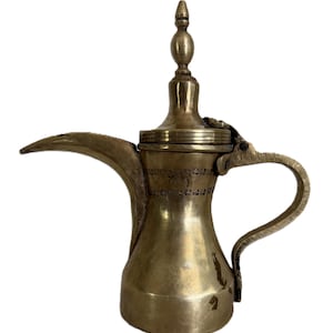 Puede incluir: Una cafetera de latón con un pico largo, un asa curvada y un adorno decorativo en la tapa. La cafetera tiene detalles grabados alrededor del cuerpo y la tapa. El diseño es ornamentado y tradicional.