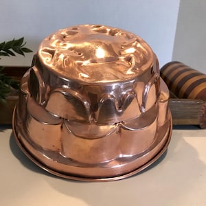 Puede incluir: Un molde de cobre para hacer un pastel o un pudín. El molde tiene un borde festoneado y un diseño en relieve de un cangrejo en la parte superior.