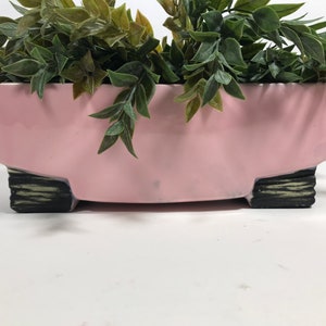 May include: A rectangular, light pink planter with a low profile, supported by four black and green textured feet. The planter is filled with green leafy faux plants, creating a decorative display. The planter is suitable for indoor or outdoor use.
